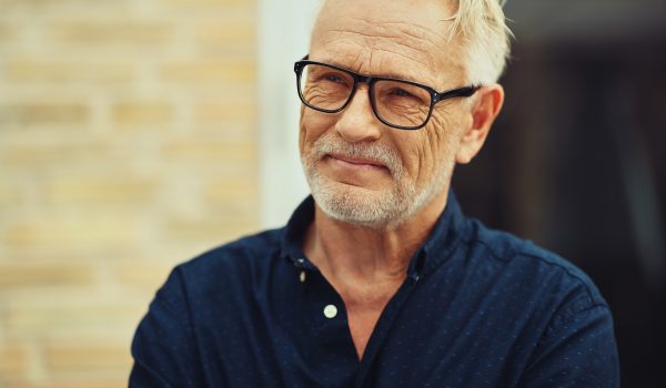 Senior man with a beard and wearing glasses smiling contently while standing by himself outside in front of his home